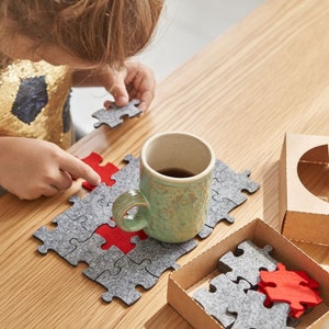 A child holding a red felt puzzle piece above a wooden table covered with grey felt puzzle pieces and the "EVADAY" box.