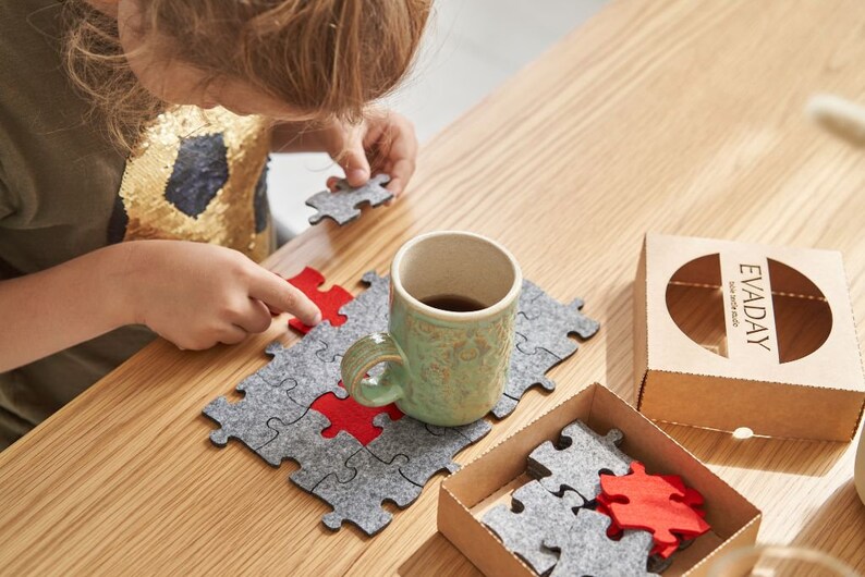 A child holding a red felt puzzle piece above a wooden table covered with grey felt puzzle pieces and the "EVADAY" box.