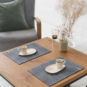 A wooden table with two grey textured placemats, white ceramic plates, and small coffee cups, alongside decorative items in a minimalist setting.