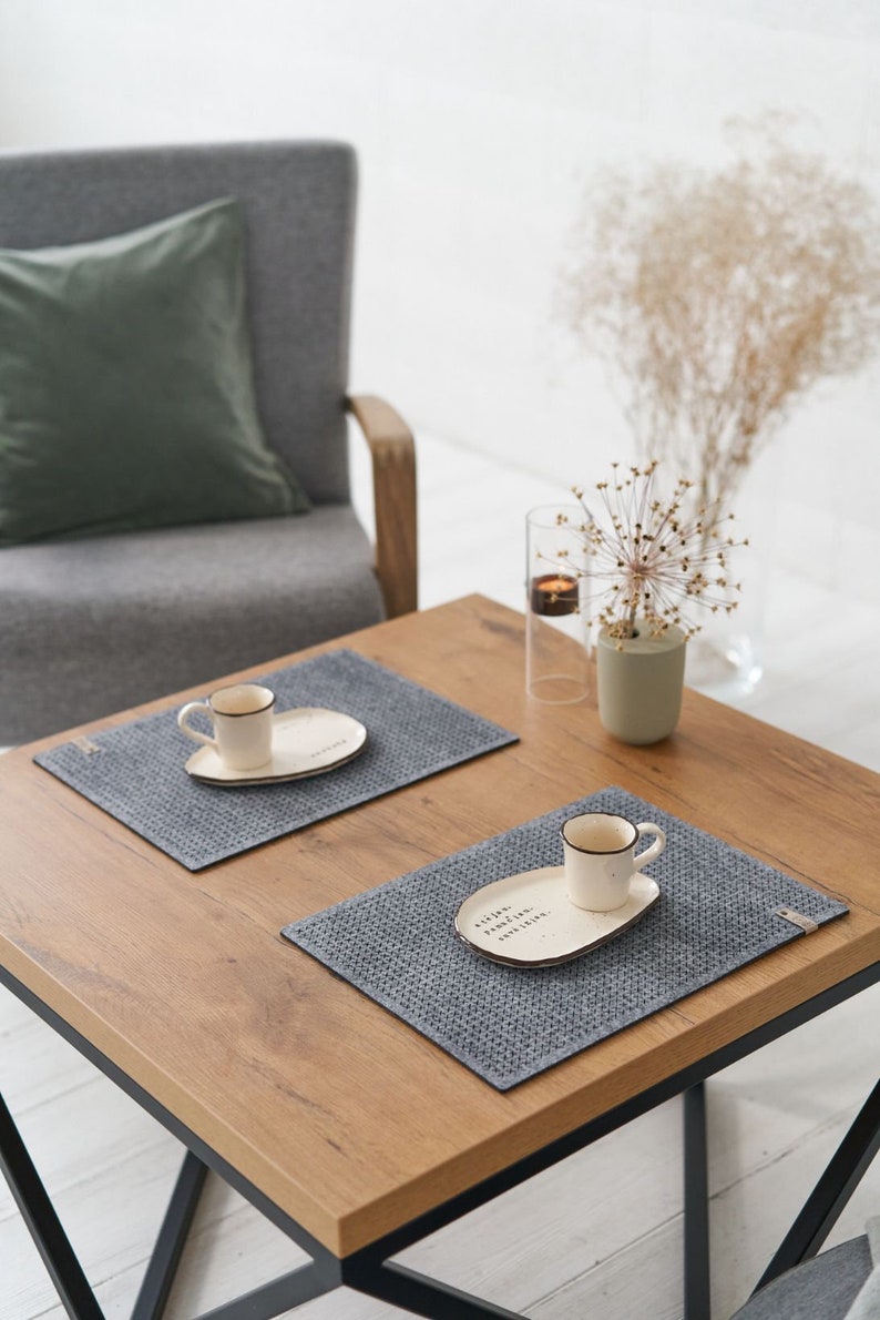 A wooden table with two grey textured placemats, white ceramic plates, and small coffee cups, alongside decorative items in a minimalist setting.