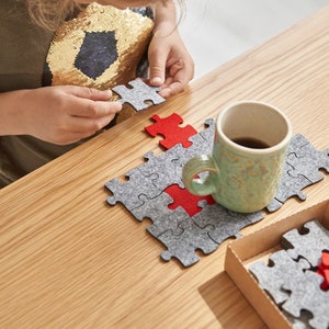 A close-up of a child's hands fitting a red felt puzzle piece into place on a wooden table with grey felt pieces.