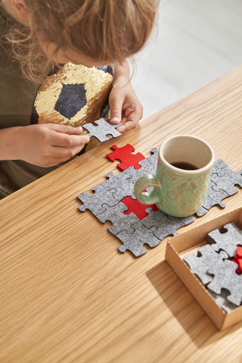 A close-up of a child's hands fitting a red felt puzzle piece into place on a wooden table with grey felt pieces.