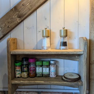 May include: A wooden shelf with two tiers, featuring a variety of spices and seasonings in glass jars. The jars are labeled with the names of the spices, such as "Garlic", "Chicken", "Chives", and "Thyme". There are also two salt and pepper shakers on the top shelf.