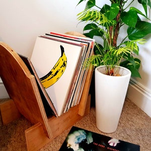 Puede incluir: Un soporte de discos de madera con una pila de discos de vinilo, incluyendo uno con un gráfico de plátano amarillo. El soporte está al lado de una maceta de cerámica blanca con una planta verde.