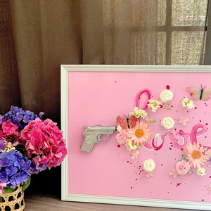 May include: A white picture frame with a pink background. The word "love" is written in pink paint with flowers and butterflies surrounding it. A silver toy gun is pointing towards the word "love".