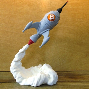 May include: A gray rocket ship figurine featuring an orange Bitcoin logo on its side. The rocket has a black pointed nose cone, three fins, and a red exhaust flame emerging from the base, which transitions into a large, white, cloud-like smoke plume.