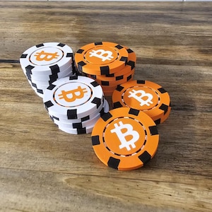 Pode incluir: Pilhas de fichas de poker Bitcoin brancas e laranja com um símbolo Bitcoin no centro. As fichas têm detalhes pretos nas bordas e estão dispostas sobre uma superfície de madeira. O símbolo Bitcoin é laranja.