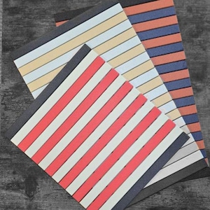 May include: Three notebooks with diagonal striped patterns in red and white, orange and blue, and cream and white. The notebooks have black and dark grey borders and are arranged on a wooden surface.