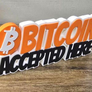 May include: A 3D sign stating "BITCOIN ACCEPTED HERE." The word "BITCOIN" is in orange and white, with the Bitcoin logo on the left. "ACCEPTED HERE" is in black. The sign is on a wooden surface.