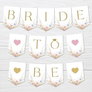 Puede incluir: Un conjunto de banderines blancos con letras doradas que dicen "BRIDE TO BE". Cada pieza del banderín está decorada con un diseño floral en colores pastel. También se incluye un anillo dorado y corazones.