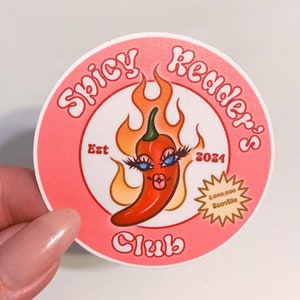 Może przedstawiać: Okrągła naklejka w kolorze różowym i białym z tekstem "Spicy Reader's Club" w retro czcionce. Naklejka przedstawia kreskówkową czerwoną papryczkę chili z rzęsami i płomieniem za nią. Na naklejce widnieje tekst "Est 2024". Na naklejce znajduje się złota gwiazdka z tekstem "2 000 000 Scoville".