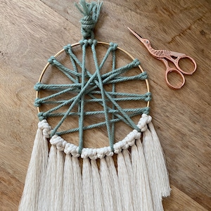 May include: A green and white macrame wall hanging with a gold hoop and tassels. The macrame is made with a thick cord and has a geometric pattern. The tassels are made with a thinner cord and are white.