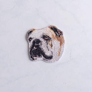 Peut inclure: Patch brodé représentant la tête d'un bouledogue. Le bouledogue a le pelage blanc avec des taches brunes autour des yeux et des oreilles. Le nez et les yeux du bouledogue sont noirs.