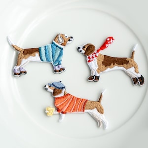 May include: Three embroidered iron-on patches of dogs wearing different outfits. The first dog is wearing a blue sweater, the second dog is wearing a red scarf, and the third dog is wearing an orange sweater.