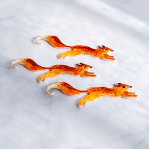 May include: Three embroidered orange and brown squirrel patches on a white background. The squirrels are in a running pose.