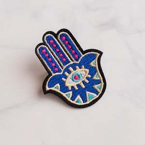 May include: Embroidered patch in the shape of a Hamsa hand, featuring a blue palm with pink and turquoise accents. The eye in the center is white with a turquoise center. The patch has a black border.