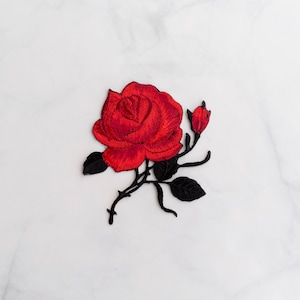May include: A red embroidered rose with black stems and leaves. The rose is a single bloom with a bud on the stem. The embroidery is on a white background.