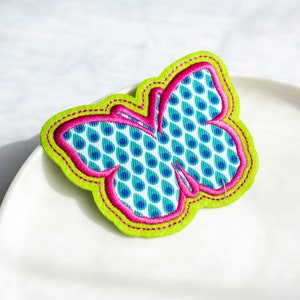 Può includere: Un'applicazione a forma di farfalla in feltro verde e rosa con un motivo a goccia blu e bianco. La farfalla è contornata da filo rosa e verde.