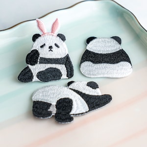 Può includere: Tre toppe ricamate a forma di panda in bianco e nero. Un panda indossa una fascia con orecchie da coniglio rosa. Le toppe sono su un piatto di ceramica azzurro e verde. I panda sono in pose diverse.