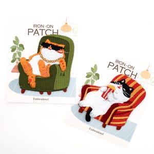 May include: Two embroidered iron-on patches. One shows an orange and white cat wearing sunglasses and a gold chain, lounging in a green armchair. The other is a black and white cat in a red striped armchair. Text reads "IRON-ON PATCH".