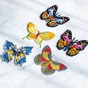 Può includere: Cinque toppe ricamate da stirare con motivi di farfalle colorate. Le farfalle sono di vari colori, tra cui blu, giallo, rosso, arancione, nero e bianco.