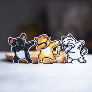Può includere: Tre toppe ricamate raffiguranti animali in posa dab. Le toppe includono un bulldog francese nero, un gatto tabby arancione con occhiali da sole e un West Highland Terrier bianco. Ogni toppa ha un bordo bianco.
