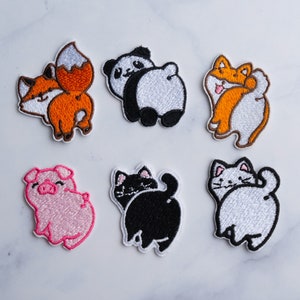 Puede incluir: Seis parches bordados para planchar que representan diferentes animales, entre ellos un zorro, un panda, un perro, un cerdo, un gato y un gato blanco. Los parches están todos de espaldas al espectador, con sus colas visibles.