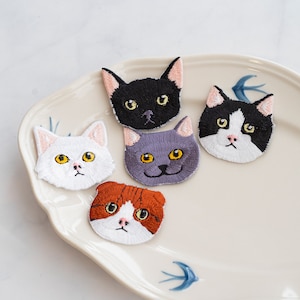 May include: Five embroidered iron-on patches featuring different cat faces. The patches are white with black, brown, gray, and white embroidery. The patches are on a white plate with a blue and white design.