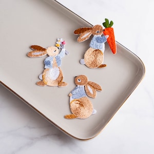May include: Three embroidered iron-on patches of brown rabbits wearing blue shirts. One rabbit holds a carrot, one holds a bouquet of flowers, and one is standing with its paws together.