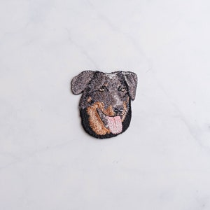 May include: Embroidered patch of a black and brown dog with a pink tongue sticking out. The dog is looking to the right.