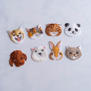 Può includere: Un set di otto toppe ricamate da stirare con diversi volti di animali, tra cui un gatto, un cane, un panda, un coniglio e altro ancora. Le toppe sono bianche con dettagli di ricamo colorati.
