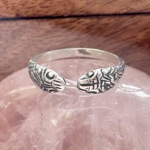 Op de afbeelding: Een zilverkleurige ring met een open ontwerp, met gedetailleerde visachtige patronen aan elk uiteinde. De ring wordt getoond op een roze, afgeronde steen, met een houten achtergrond. De ring is een sieraad.