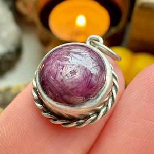 May include: A circular silver pendant featuring a large, polished, deep-red gemstone. The gemstone is set within a twisted silver rope design. The pendant is held between fingers, with a blurred background including a candle and other objects.