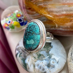 May include: A silver ring with a turquoise stone set in a bezel setting. The stone has a green and blue color with a marbled pattern.