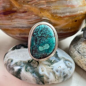 May include: A silver ring with a turquoise stone set in a bezel setting. The stone has a green and blue color with black veins running through it.