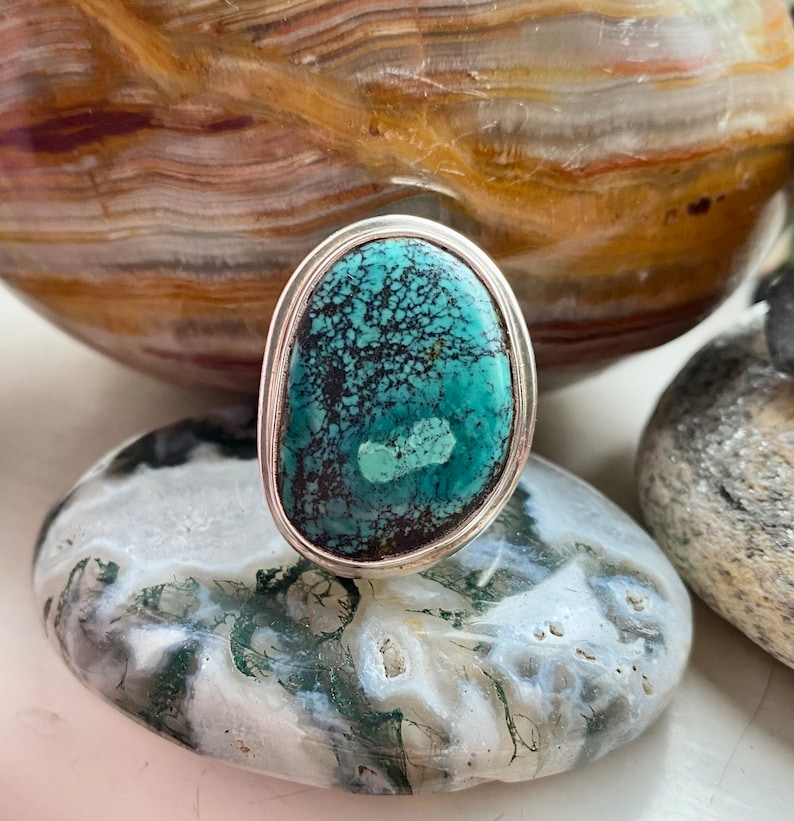 May include: A silver ring with a turquoise stone set in a bezel setting. The stone has a green and blue color with black veins running through it.