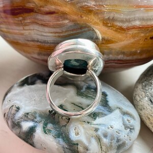 May include: A silver ring with a green gemstone set in a bezel setting. The ring is on a white and green stone background.
