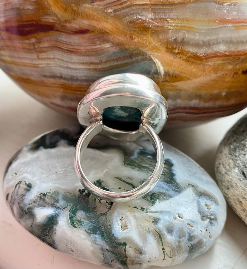 May include: A silver ring with a green gemstone set in a bezel setting. The ring is on a white and green stone background.