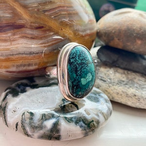May include: A silver ring with a large oval turquoise stone set in a bezel setting. The stone has a dark green and black matrix.