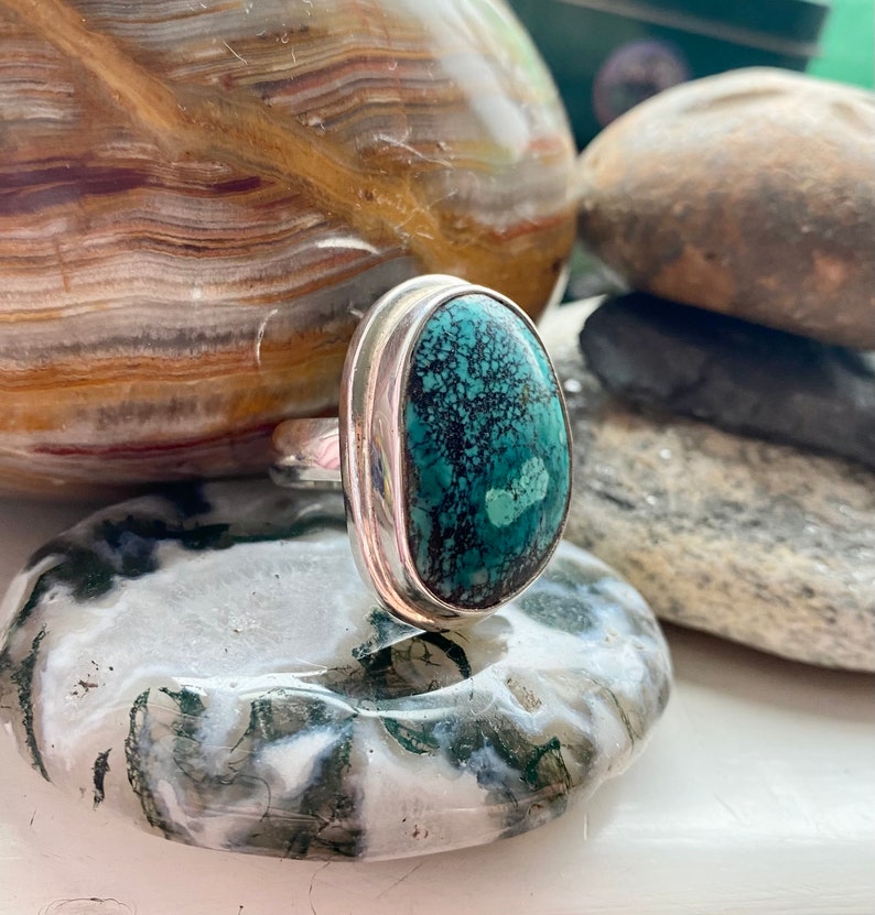 May include: A silver ring with a large oval turquoise stone set in a bezel setting. The stone has a dark green and black matrix.