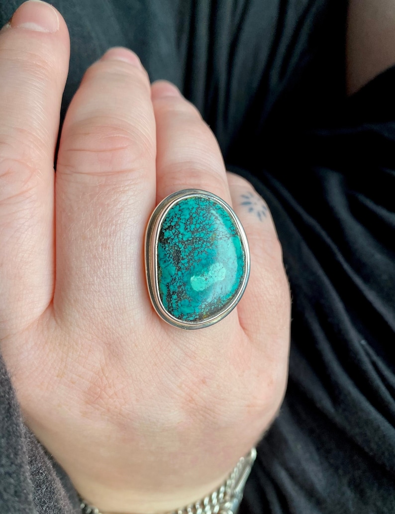 May include: A silver ring with a large oval turquoise stone set in a bezel setting. The stone has a dark green color with black veins running through it.