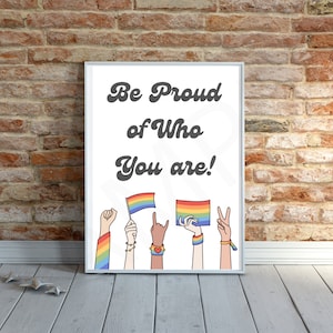 Puede incluir: Impresión enmarcada en blanco con el texto "Be Proud of Who You Are!" en fuente cursiva negra. La impresión presenta una bandera arcoíris y varias manos sosteniendo banderas arcoíris y pulseras.