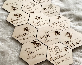 May include: Wooden hexagonal baby milestone cards with a bee and honeycomb design. The cards feature the numbers one through eleven and one year, with the words "months" or "year" written below each number.