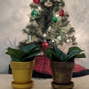 May include: Two small, decorative flower pots with faux green plants. One pot is gold, the other is brown with gold accents. Both pots have matching saucers. A Christmas tree with ornaments is in the background.