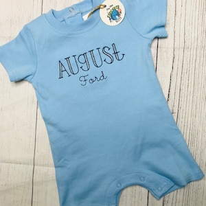 May include: Light blue baby onesie with short sleeves and snaps at the bottom. The onesie has the name "AUGUST Ford" embroidered on the front in navy blue thread.