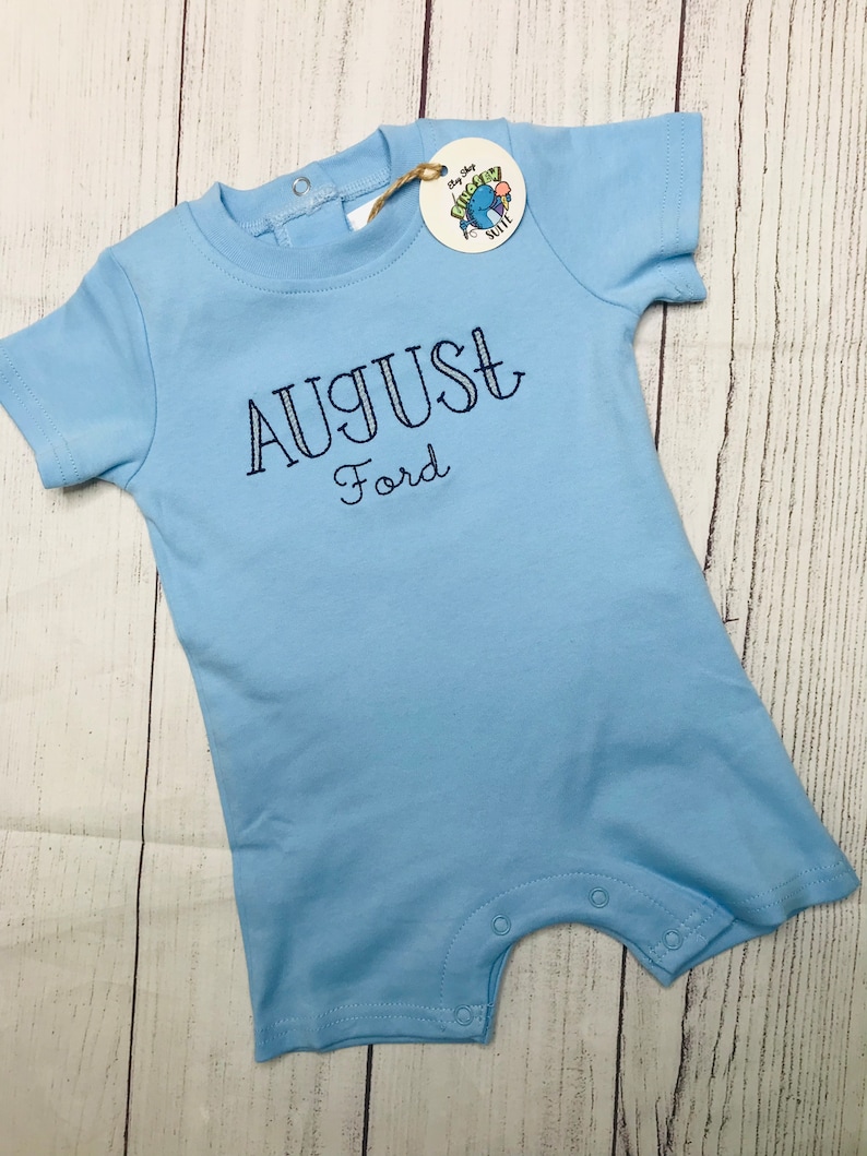 May include: Light blue baby onesie with short sleeves and snaps at the bottom. The onesie has the name "AUGUST Ford" embroidered on the front in navy blue thread.