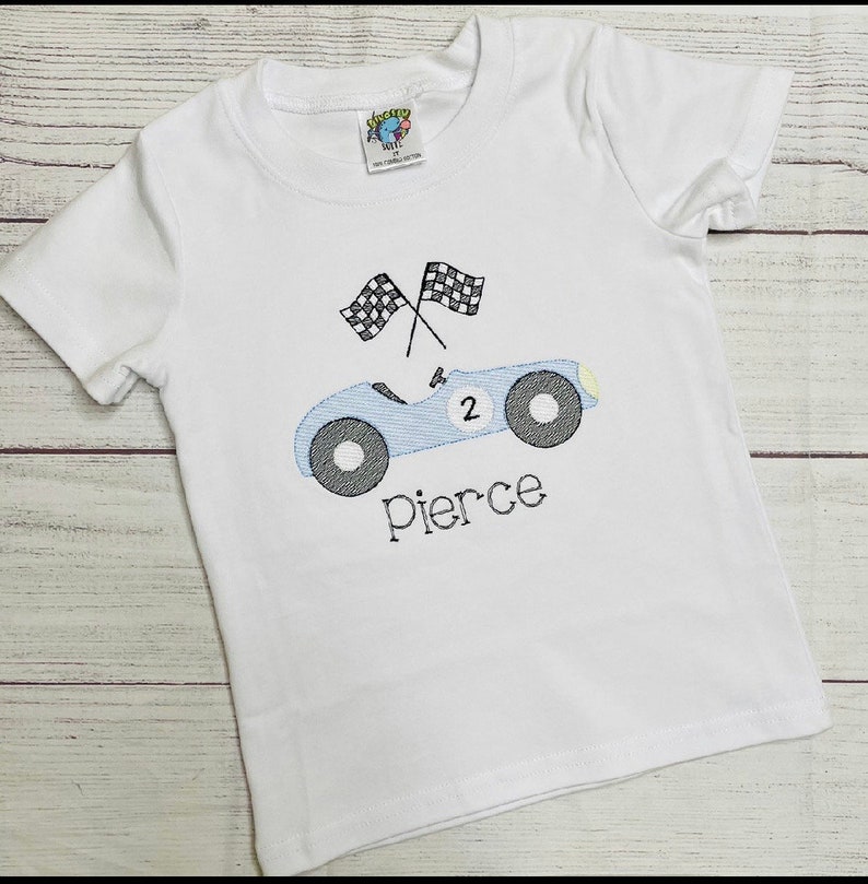 Peut inclure: T-shirt blanc &agrave; manches courtes avec un motif de voiture de course bleu clair et des drapeaux &agrave; damier noir et blanc. La voiture porte le num&eacute;ro 2 et le nom "Pierce" brod&eacute; en dessous. Le t-shirt a une &eacute;tiquette en haut &agrave; gauche.