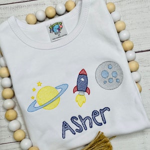 May include: White baby onesie with an embroidered design of a rocket, planet, and moon. The name "Asher" is embroidered in blue below the design.