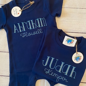 May include: Two navy blue baby onesies with embroidered names. The onesie on the left says "Abraham Howell" and the onesie on the right says "Judah Kemper".