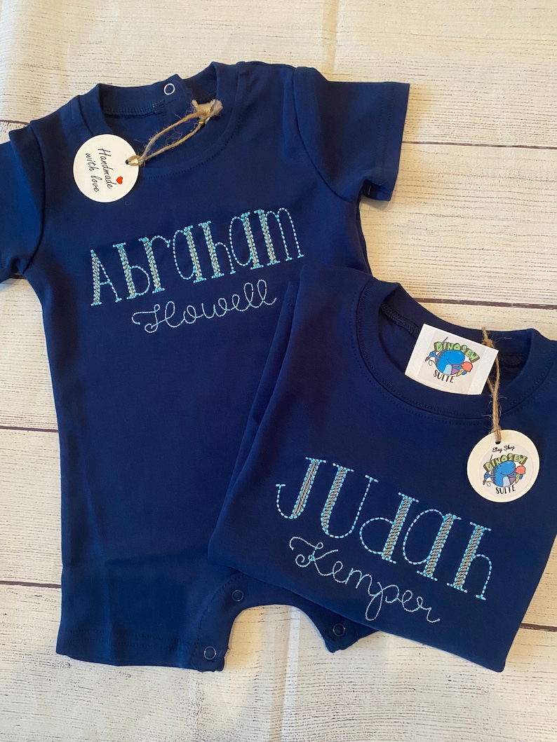 May include: Two navy blue baby onesies with embroidered names. The onesie on the left says "Abraham Howell" and the onesie on the right says "Judah Kemper".
